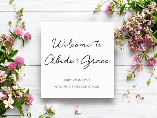 Flat lay of a white square card on a whitewashed wood background, surrounded by blush and pink flowers. The card reads “Welcome to Abide & Grace” in elegant script, with the tagline “Abiding in love. Creating through grace.” beneath it.